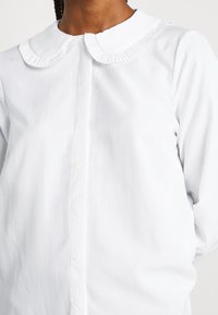 Camicia bianca con colletto rotondo con bordo plissettato, chiusura frontale con bottoni, maniche lunghe e tessuto liscio.
