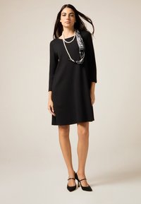 Czarna sukienka z rękawami do łokci, o luźnym kroju i długości do kolan. Uzupełniona wzorzystą apaszką i czarnymi butami na obcasie.