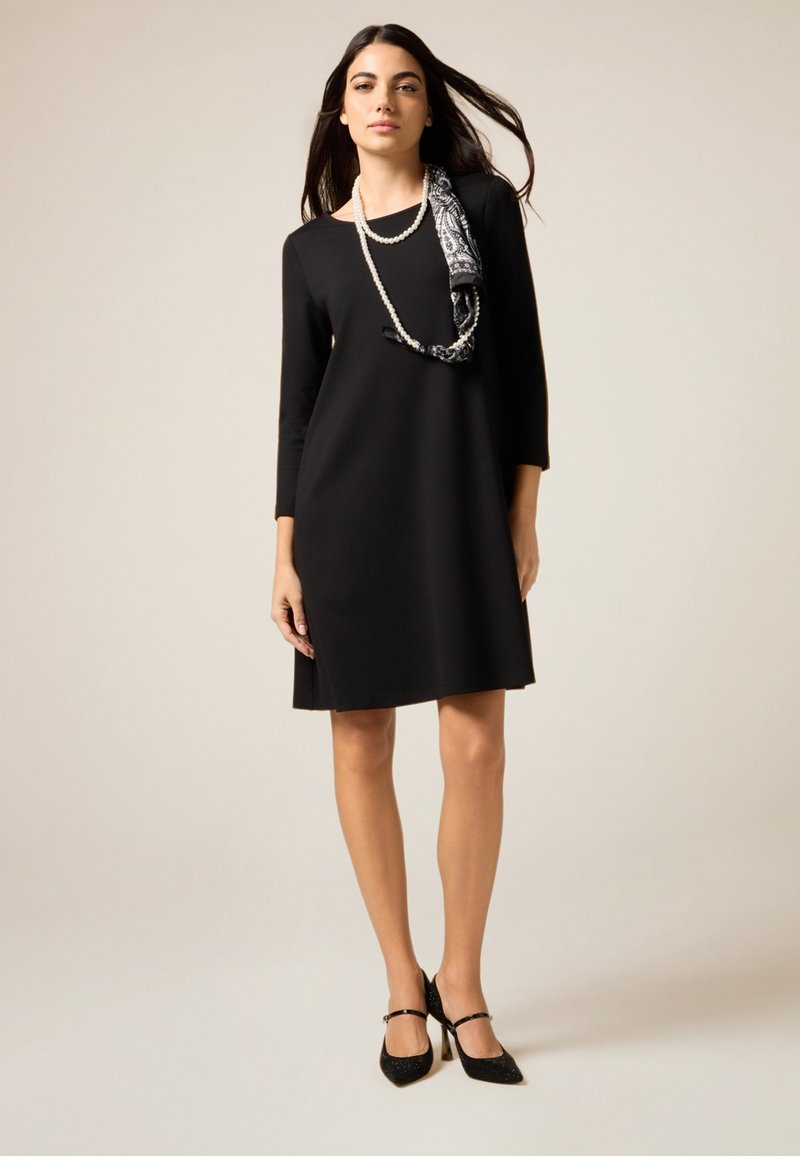 Czarna sukienka z rękawami do łokci, o luźnym kroju i długości do kolan. Uzupełniona wzorzystą apaszką i czarnymi butami na obcasie.