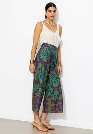 Femme portant un haut tricoté sans manches blanc, un pantalon taille haute à jambes larges avec des motifs floraux verts et violets, des bracelets dorés et des sandales.