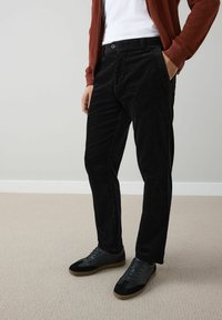 Black corduroy pants with a slim-fit design. Features front pockets and a button closure. Paired with black sneakers and a rust-colored jacket.