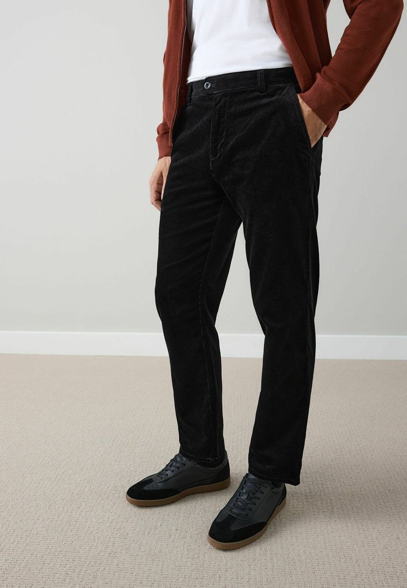 Black corduroy pants with a slim-fit design. Features front pockets and a button closure. Paired with black sneakers and a rust-colored jacket.