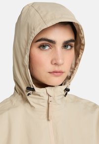 Beige huvjacka med en slät textur, justerbara snoddar och en fullständig frontdragkedja. Har en minimalistisk design utan mönster.