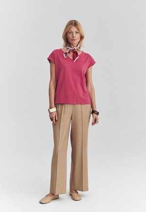 Mujer de pie con una camiseta rosa de manga corta, pantalones anchos beige, zapatos planos beige, pañuelo con estampado en el cuello y pulseras en ambas muñecas.