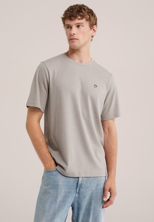 Jeune homme aux cheveux bouclés châtain clair portant un t-shirt uni gris clair à manches courtes et un jean bleu sur un fond neutre.