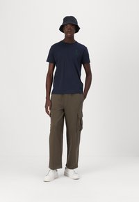 Navy cotton T-shirt with green number print, olive cargo trousers, and white sneakers. The model wears a black bucket hat.