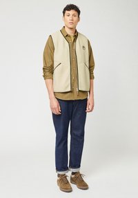 Gilet in pile beige con chiusura a zip, indossato sopra una camicia verde oliva a bottoni. Jeans in denim scuro con risvolti e stivali marroni con lacci.