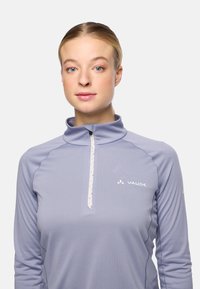 Junge Frau mit heller Haut und nach hinten gebundenem blonden Haar, trägt ein hellviolettes VAUDE-Sportoberteil mit Reißverschluss und steht mit Blick zur Kamera vor einem schlichten Hintergrund.
