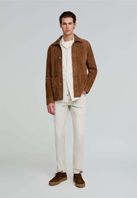 Braune Wildlederjacke mit einem Hemd darunter, hellen Hosen und braunen Schuhen. Die Jacke verfügt über einen Kragen und zwei Fronttaschen.