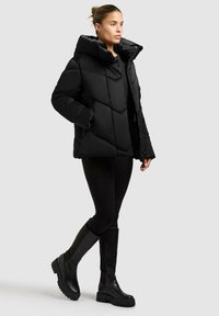 Schwarze Daunenjacke mit hohem Kragen, ausgestattet mit geometrischem Steppmuster und seitlichen Taschen, getragen über schwarzen Leggings und Knielangen Stiefeln.