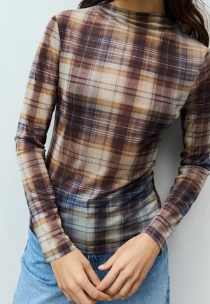 Femme portant un haut transparent ajusté à manches longues avec un motif à carreaux marron, beige et gris, associé à un jean bleu clair.