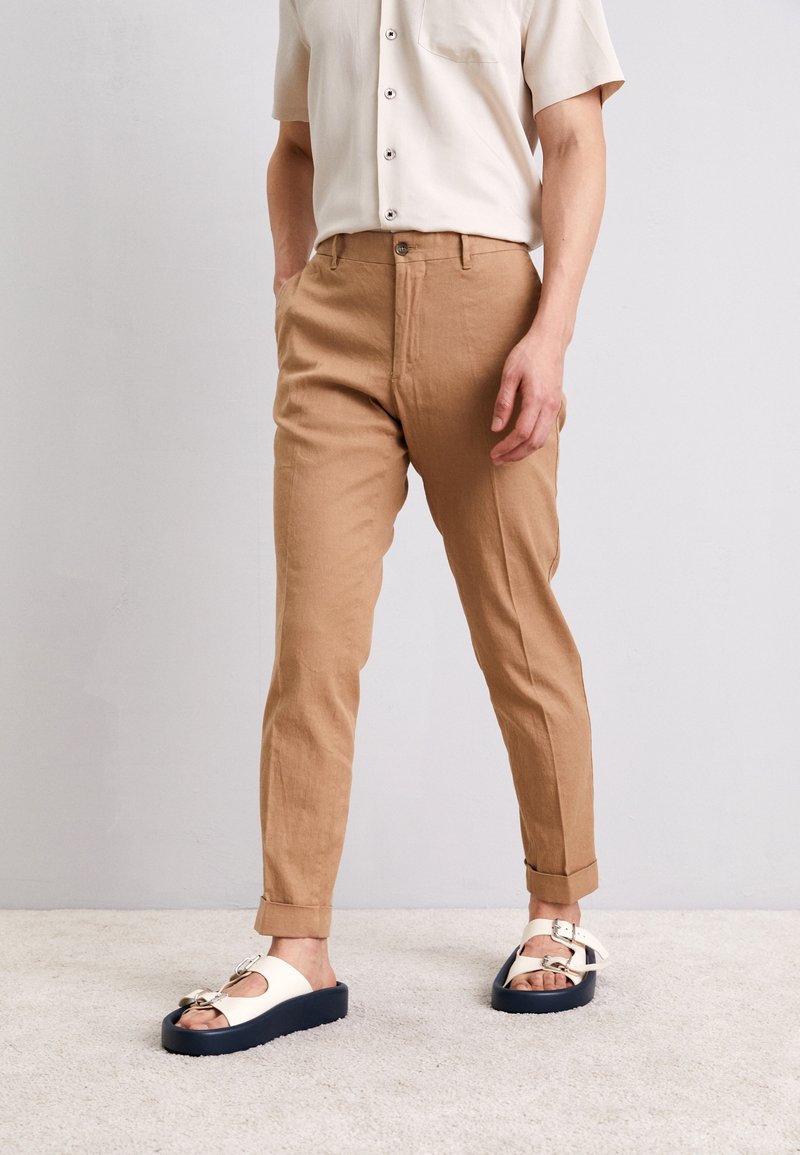 Bruna, avsmalnande byxor med veckad front, uppvikta fållar och en slät textur. I kombination med vita sandaler med mörk sula och silverdetaljer.