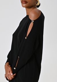 Donna che indossa un maglione nero con ritagli sulle spalle fissati da bottoni argentati, mentre tiene la manica con entrambe le mani.