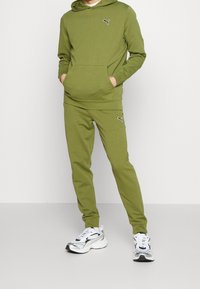 Man klädd i en olivgrön Puma träningsoverall med hoodie och joggers, tillsammans med vita och silverfärgade Puma sneakers, stående mot en neutral bakgrund.