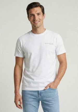 Glunderende man die een wit T-shirt met korte mouwen draagt met kleine tekst op de borst en lichtblauwe jeans, staand met één hand in de zak.