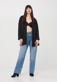 Cardigan lungo nero con maniche lunghe, aperto e dotato di tasche laterali, indossato sopra un reggiseno in pizzo nero e jeans in denim azzurro chiaro a vita alta, caratterizzati da una linea dritta.