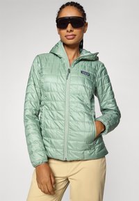 Woman wearing dark sunglasses and a light green quilted jacket with hands in pockets, standing against a plain background.