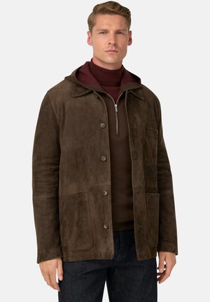 Man in een bruine suède jack over een kastanjebruine kwart-rits trui, gecombineerd met donkere jeans, staand tegen een effen achtergrond.
