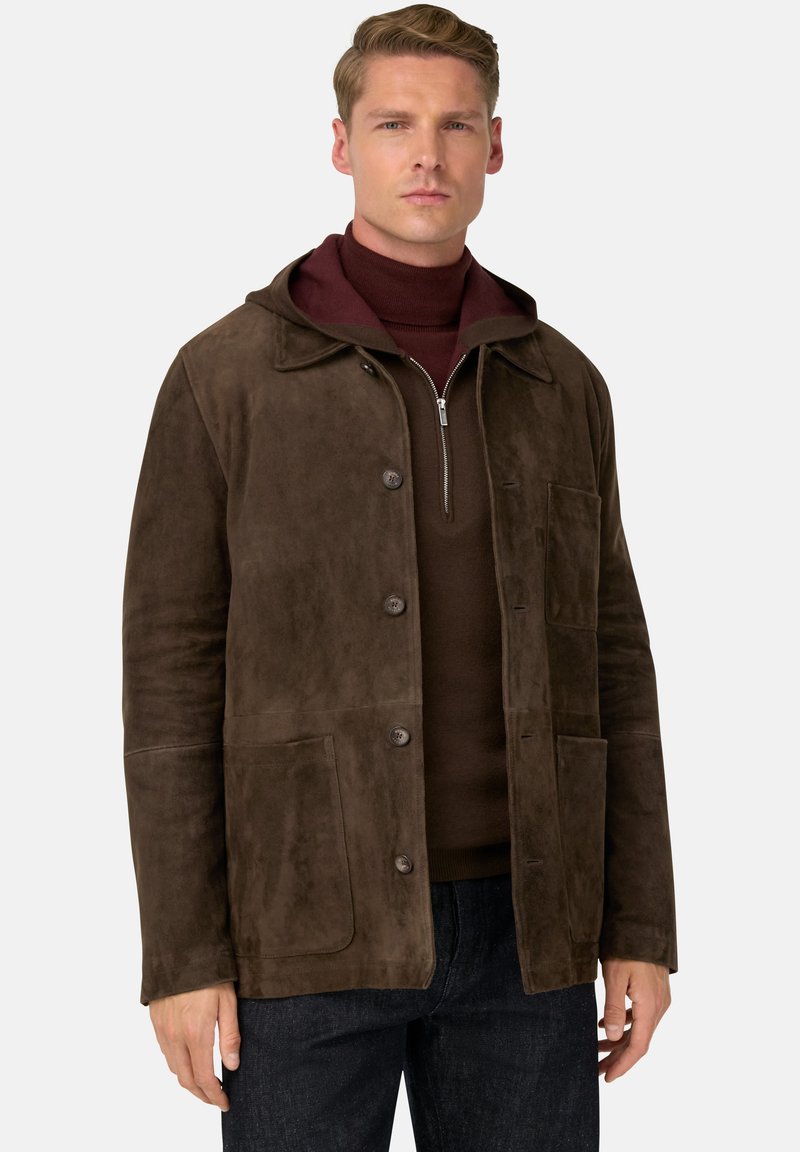 Man som bär en brun mockajacka över en vinröd tröja med halv dragkedja, tillsammans med mörka jeans, står mot en neutral bakgrund.