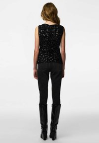 Svart paljettbesatt ermeløs topp kombinert med svarte høytlivsjeans og knelangs svarte skinnstøvler, sett bakfra.