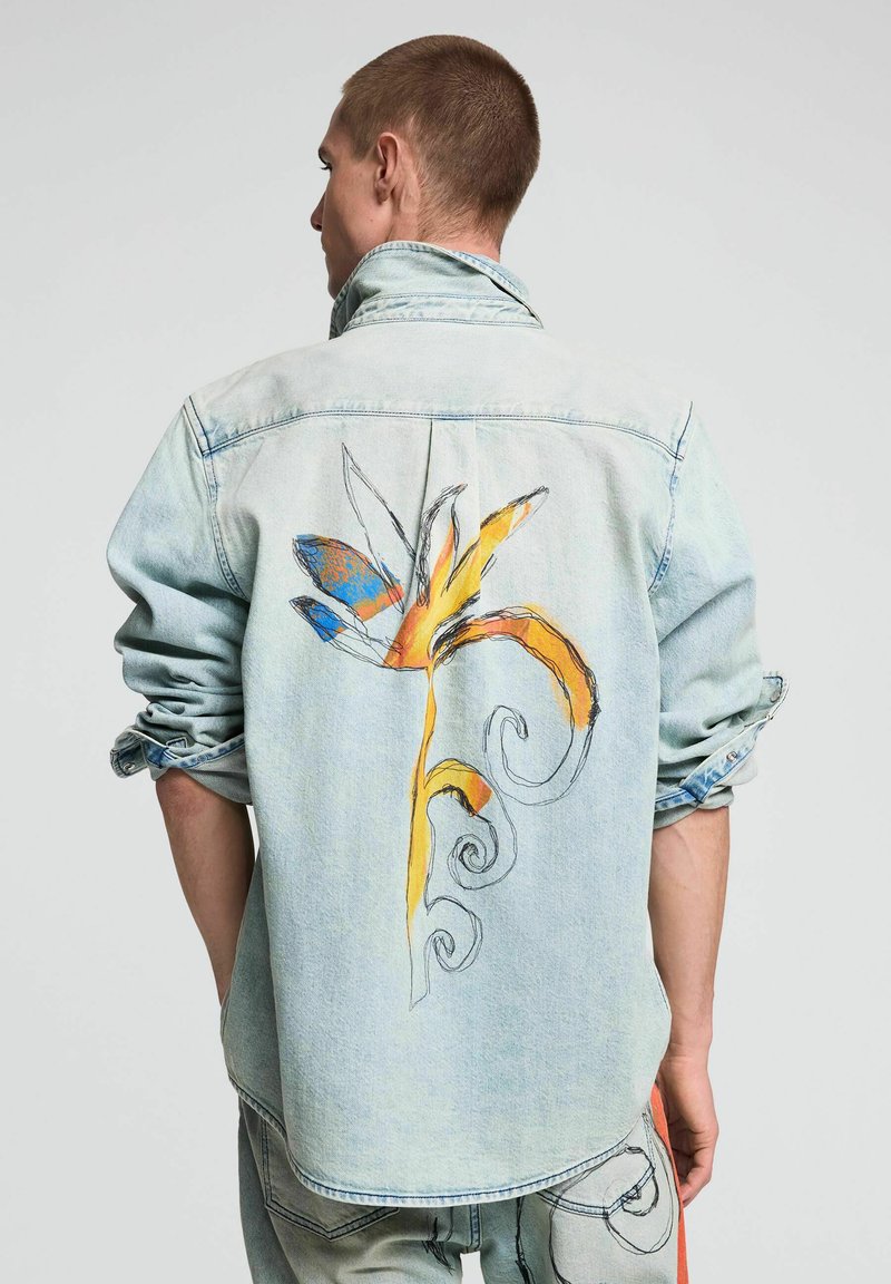 Hombre de cabello corto lleva una chaqueta vaquera azul claro con diseño floral abstracto y colorido en la espalda, mangas arremangadas.