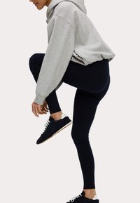 Grijze hoodie, zwarte leggings en zwarte sneakers met witte accenten. De materialen lijken zacht te zijn en van suède. De leggings zijn aansluitend.
