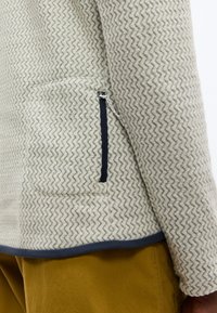 Φλις μπουφάν μπεζ με σχέδιο ζιγκ-ζαγκ, με ναυτικό τελείωμα και τσέπη με φερμουάρ, φορεμένο πάνω από παντελόνι μουσταρδί χρώματος.