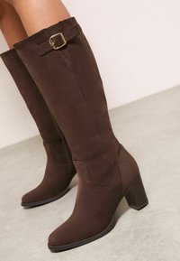 Botas altas hasta la rodilla de ante marrón con punta afilada, tacón grueso y un adorno decorativo con hebilla en la parte superior. Textura suave.