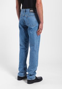 Modré džínové kalhoty s rovnými nohavicemi, pěti kapsami a černou značkovou etiketou na pasu. Nahoře s černými botami, viděno zezadu.