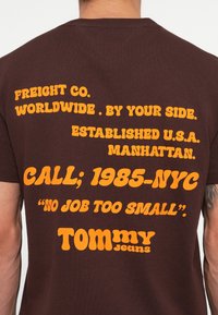 Tricou din bumbac maro, având text portocalie îndrăzneț care afișează numele brandului, sloganul și informațiile de contact, cu o croială lejeră și mâneci scurte.
