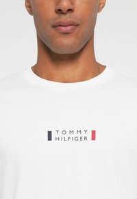 Bijela pamučna majica s logotipom "TOMMY HILFIGER" u crnoj boji, s crvenim i mornarsko plavim akcentima, okruglim izrezom i kratkim rukavima.