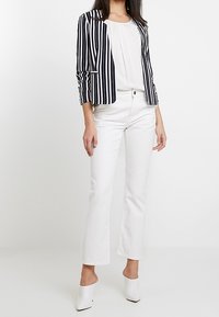 Blazer rayé bleu marine et blanc, blouse blanche, et jean évasé blanc. Mules blanches à talons hauts. Tissu à texture légère, coupe ajustée.