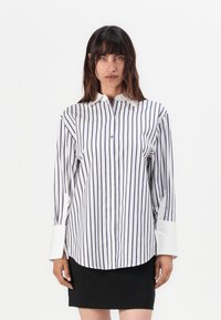 Chemise rayée blanche et bleu marine avec un col pointu, des manches longues et un ourlet arrondi. Elle présente des boutons de fermeture et des poignets blancs contrastants.