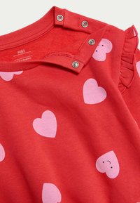 Rød bomulds t-shirt med korte ærmer, dekoreret med lyserøde hjerte-mønstre og en lille flæse-detalje. Har trykknaplukninger ved skulderen for nem påklædning.