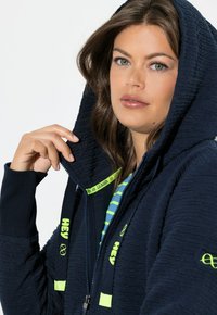 Tmavě modrá kapsáčová bunda s texturovanou tkaninou, s neonově žlutými akcenty, zapínáním na zip a šňůrkami s logem značky.