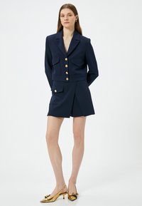 Marineblauwe blazer met gouden knopen, voorzien van passende zakken en een getailleerd ontwerp, gecombineerd met bijpassende marineblauwe shorts en glanzende gouden hakken.