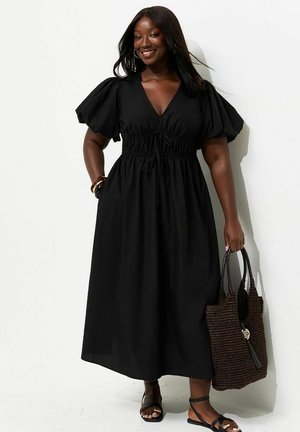 Uśmiechnięta kobieta w czarnej sukience midi z bufkami na rękawach i sandałach, trzymająca dużą plecioną torbę, stojąca na tle białego tła.