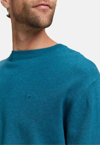 Blauer Strickpullover mit Rundhalsausschnitt, gerippten Bündchen und einem kleinen Logodetail auf der Brust. Weiche Textur mit einer körpernahen Silhouette.