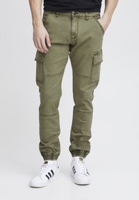 Pantalons cargo en vert olive, dotés de plusieurs poches, de revers fuselés, d'une fermeture à bouton et d'un tissu doux et texturé. Associés à des baskets blanches.