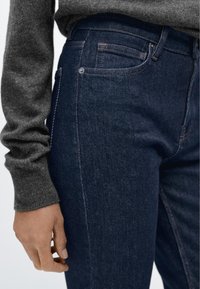 Mörkblå jeans med smal passform, utrustade med klassisk design med fem fickor, synliga sömmar och en subtil textur. Grå tröjärm syns delvis.