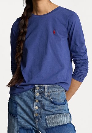 Persoon draagt een blauw shirt met lange mouwen en een rood Polo-logo, hoge patchwork spijkerbroek met knoopsluiting en gevlochten haar.