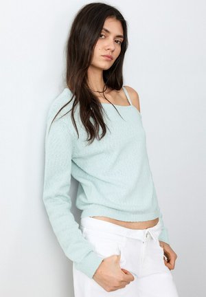 Jeune femme aux longs cheveux foncés portant un pull bleu clair à épaules dénudées et un pantalon blanc, appuyée contre un mur blanc.