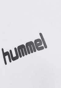 Tessuto bianco con un logo nero prominente riportante il testo "hummel" in un font arrotondato e in grassetto. La texture appare liscia ed elastica.