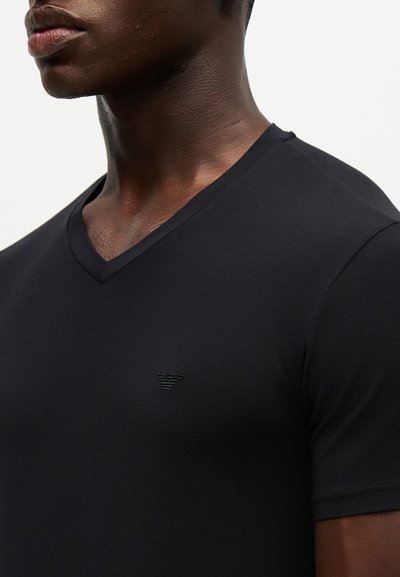 Άνδρας που φοράει μαύρο T-shirt με V-λαιμόκοψη και μικρό τονικό λογότυπο αριστερά στο στήθος, εμφανίζεται από τον λαιμό έως το πάνω μέρος του κορμού.