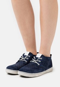 Scarpe scamosciate blu navy con motivo geometrico perforato, lacci bianchi e suola in gomma. Presentano un colletto imbottito e una finitura testurizzata.