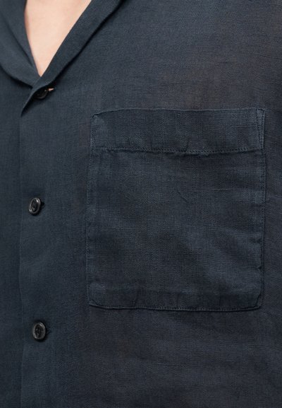 Σκούρο μπλε λινό πουκάμισο με κλείσιμο με κουμπιά μπροστά και τσέπη στο στήθος. Το ύφασμα έχει υφή με ελαφριά γυαλάδα.