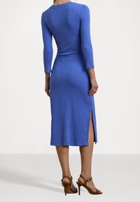 Långärmad, blå, åtsittande klänning med midikjol och sidofickor. Slät textur. Bärs med bruna högklackade sandaler.