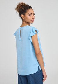 Top azzurro chiaro a maniche corte con dettagli arricciati, scollo rotondo e apertura a chiave sul retro. Realizzato in un tessuto morbido e liscio.