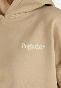 Bežinė gobtuvinė palaidinė, pagaminta iš minkštos medžiagos, su išsiuvinėtu "Pegador" logotipu baltos spalvos, turinti lygią tekstūrą ir siuvinėjimo detales.