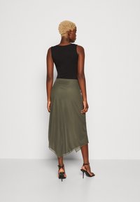Black sleeveless top with a high neckline, paired with a green asymmetric skirt; model wears black high-heeled sandals, showing smooth textures.